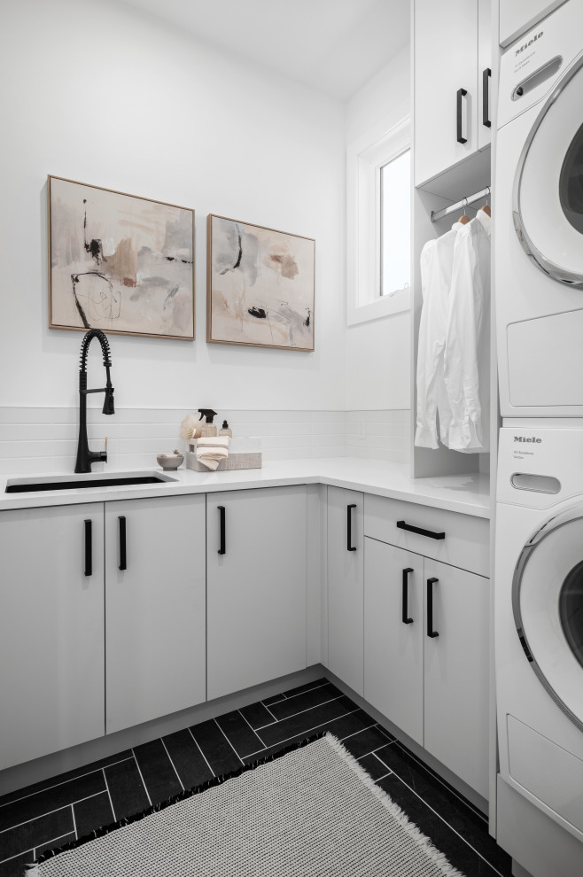This is an example of a mid-sized modern l-shaped dedicated laundry room in Calgary with an undermount sink, flat-panel cabinets, grey cabinets, quartz benchtops, grey splashback, ceramic splashback, white walls, porcelain floors, a stacked washer and dryer, grey floor and white benchtop.