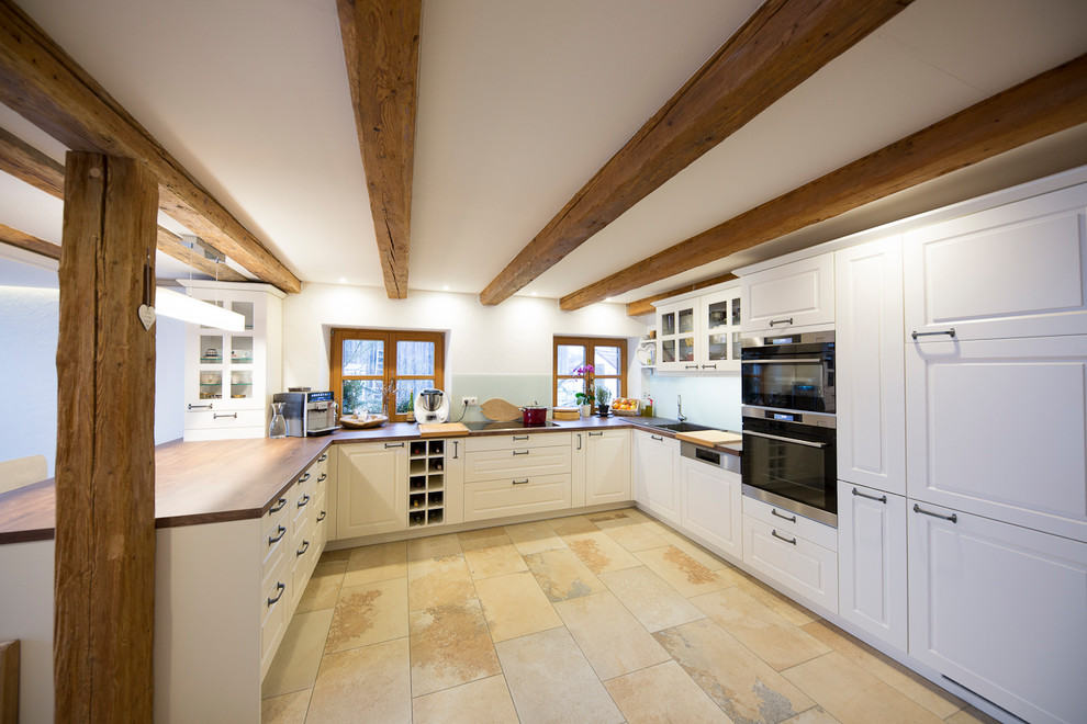 Photo of a country u-shaped kitchen in Munich with shaker cabinets, white cabinets, wood benchtops, black appliances, a peninsula, beige floor, brown benchtop, a drop-in sink, blue splashback, glass sheet splashback and cement tiles.