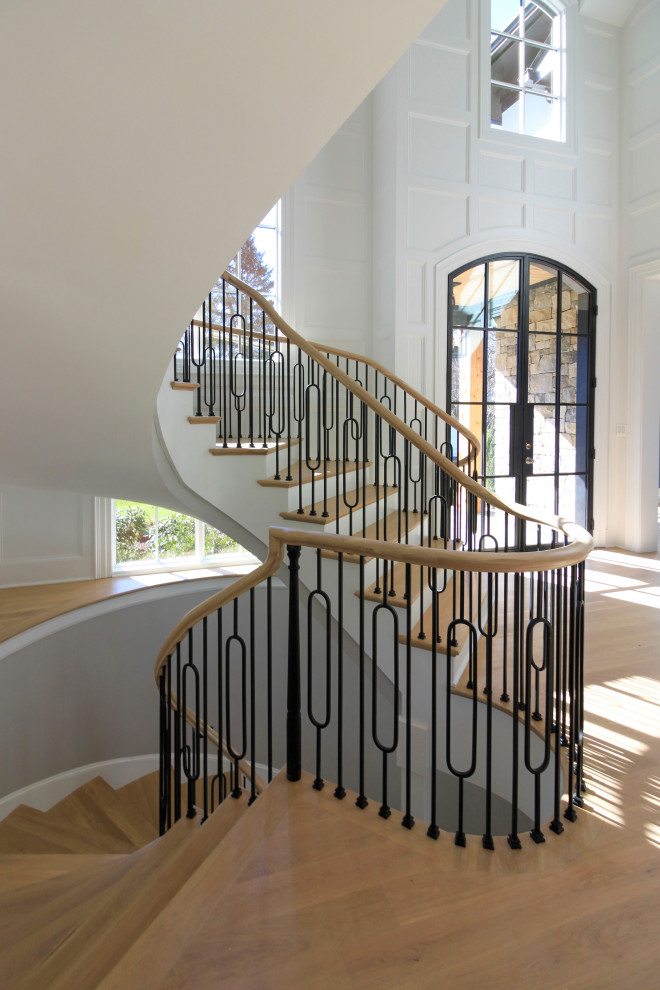 Стильный дизайн: огромная изогнутая деревянная лестница в стиле неоклассика (современная классика) с деревянными ступенями, перилами из смешанных материалов и панелями на стенах - последний тренд
