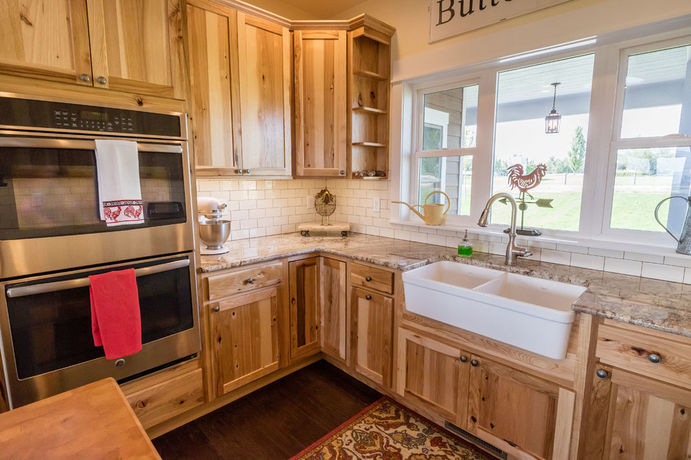 This is an example of a mid-sized country kitchen in Other with a farmhouse sink, shaker cabinets, light wood cabinets, granite benchtops, white splashback, ceramic splashback, stainless steel appliances, dark hardwood floors, with island and brown floor.