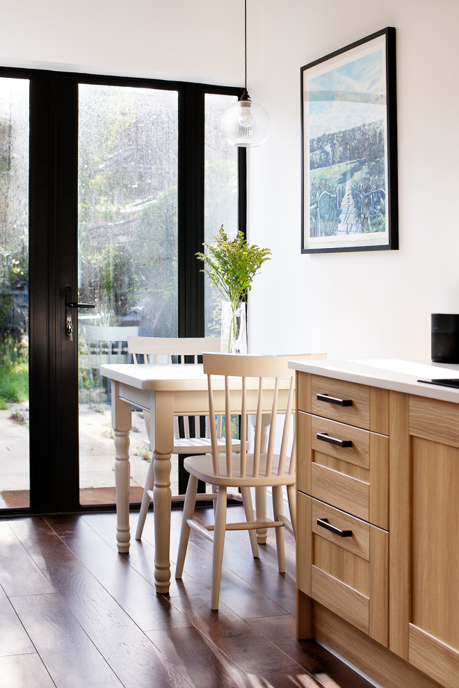 This is an example of a mid-sized contemporary galley eat-in kitchen in London with shaker cabinets, light wood cabinets, solid surface benchtops, white splashback, dark hardwood floors, no island, brown floor and white benchtop.