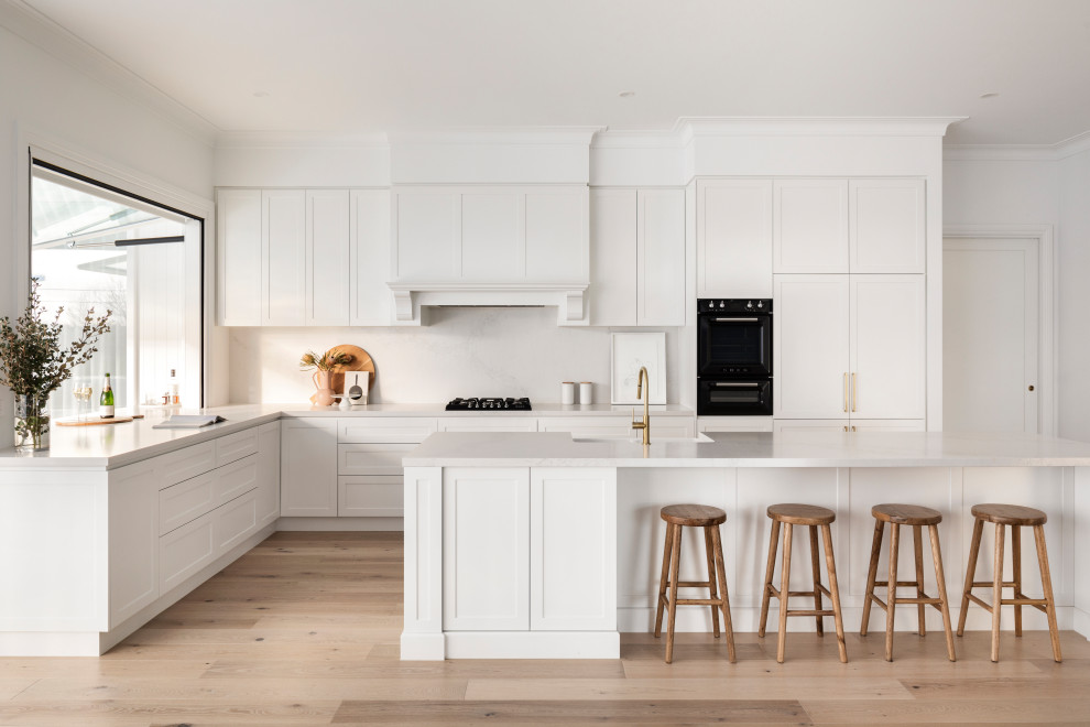Transitional l-shaped kitchen in Melbourne with a farmhouse sink, shaker cabinets, white cabinets, white splashback, black appliances, light hardwood floors, with island, beige floor and white benchtop.