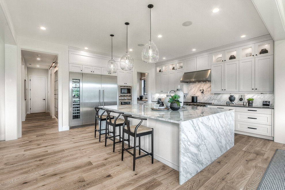 Photo of an expansive transitional l-shaped open plan kitchen in Seattle with recessed-panel cabinets, white cabinets, stainless steel appliances, light hardwood floors, with island, grey benchtop and an undermount sink.