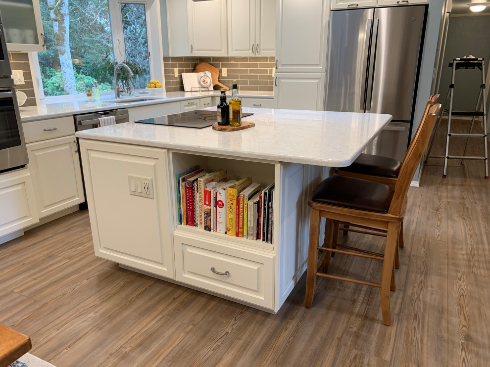 Inredning av ett klassiskt mellanstort vit vitt kök, med en undermonterad diskho, luckor med upphöjd panel, vita skåp, bänkskiva i kvarts, brunt stänkskydd, stänkskydd i tunnelbanekakel, rostfria vitvaror, laminatgolv, en köksö och brunt golv