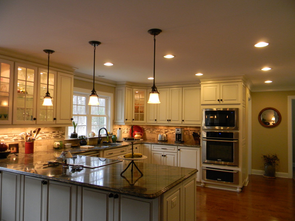 Photo of a mid-sized arts and crafts u-shaped eat-in kitchen in Charlotte with white cabinets, medium hardwood floors, a double-bowl sink, raised-panel cabinets, granite benchtops, beige splashback, stone tile splashback, stainless steel appliances and a peninsula.