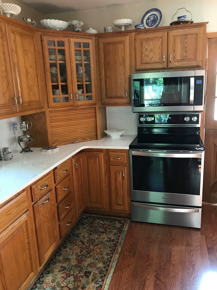 This is an example of a small traditional l-shaped separate kitchen in Cedar Rapids with an undermount sink, raised-panel cabinets, medium wood cabinets, quartz benchtops, white splashback, subway tile splashback, stainless steel appliances, carpet, no island, multi-coloured floor and white benchtop.