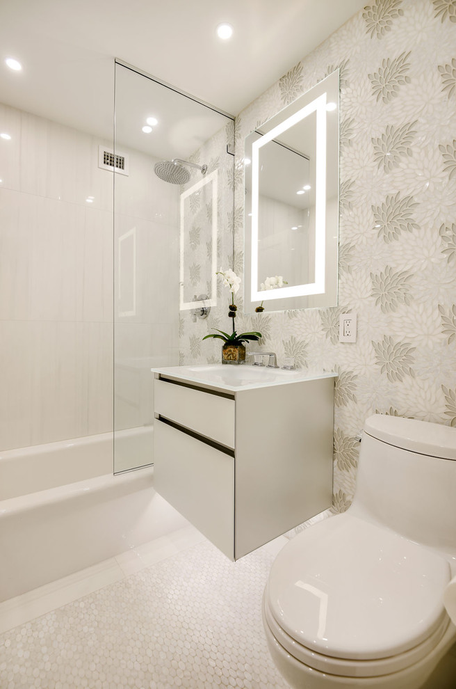 Design ideas for a small transitional bathroom in New York with glass-front cabinets, grey cabinets, an alcove tub, a shower/bathtub combo, a one-piece toilet, gray tile, ceramic tile, grey walls, ceramic floors, an integrated sink, glass benchtops, grey floor, a hinged shower door and white benchtops.