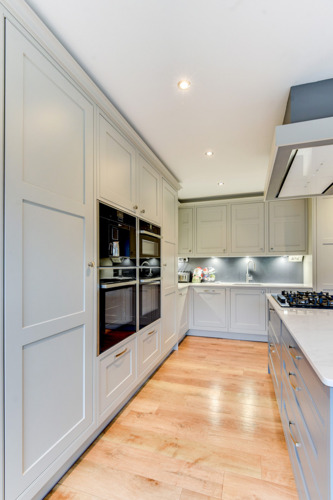 Mid-sized traditional l-shaped eat-in kitchen in Sussex with an undermount sink, shaker cabinets, grey cabinets, quartzite benchtops, grey splashback, glass sheet splashback, panelled appliances, with island, brown floor and white benchtop.
