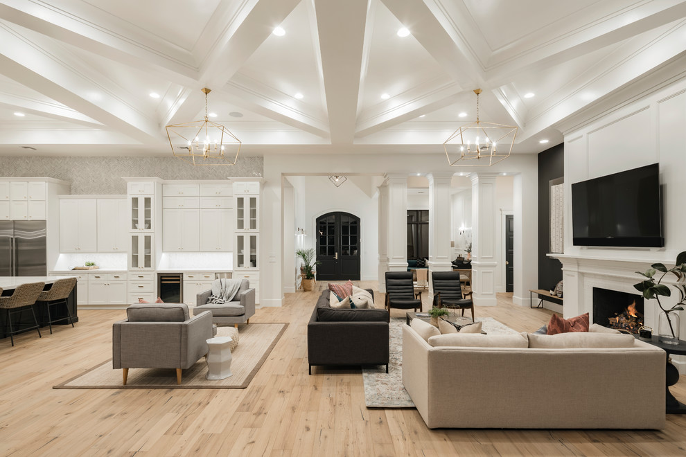 Photo of an expansive transitional open concept family room in Phoenix with a game room, white walls, light hardwood floors, a standard fireplace, a wood fireplace surround, a wall-mounted tv and beige floor.