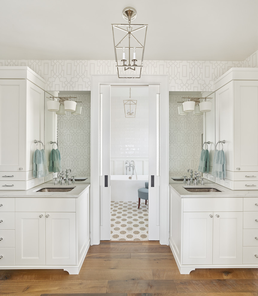 Photo of a beach style master bathroom in Charleston with recessed-panel cabinets, white cabinets, a freestanding tub, white walls, medium hardwood floors, an undermount sink, brown floor and grey benchtops.