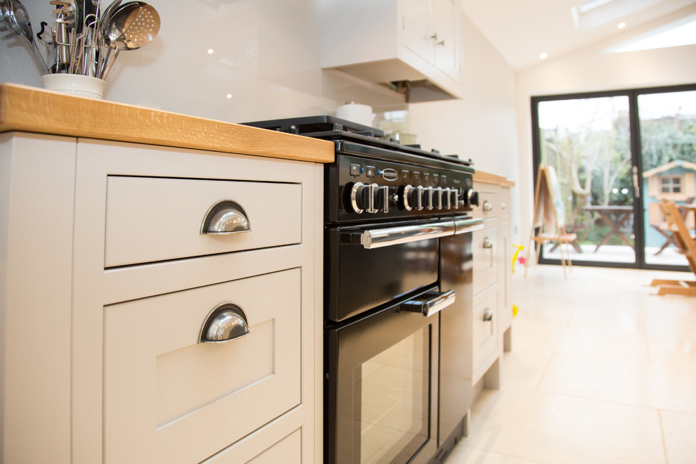 Photo of a mid-sized modern galley eat-in kitchen in London with a farmhouse sink, shaker cabinets, grey cabinets, wood benchtops, grey splashback, glass sheet splashback, black appliances, ceramic floors, with island, beige floor and brown benchtop.