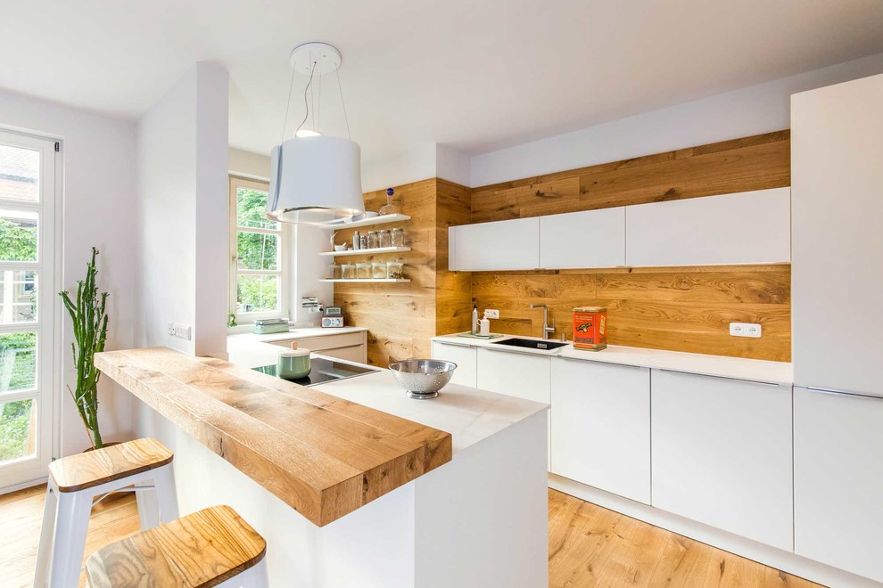 Inspiration for a mid-sized scandinavian u-shaped open plan kitchen in Nuremberg with an integrated sink, flat-panel cabinets, white cabinets, brown splashback, timber splashback, medium hardwood floors, a peninsula, brown floor and white benchtop.