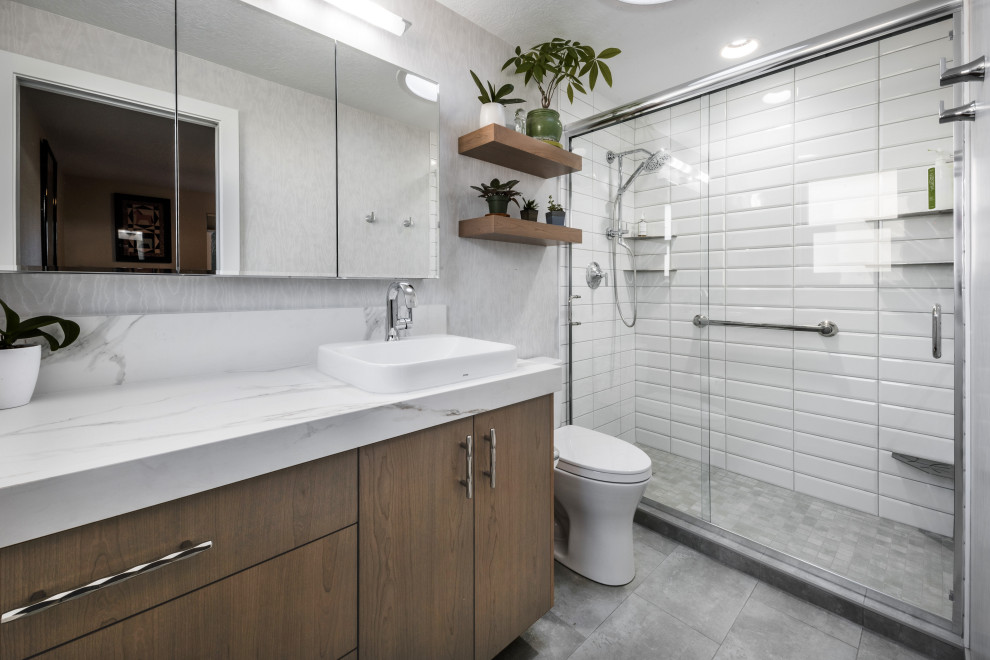 This is an example of a mid-sized contemporary master bathroom in Boise with flat-panel cabinets, medium wood cabinets, an alcove shower, a two-piece toilet, white tile, porcelain tile, grey walls, porcelain floors, a vessel sink, grey floor, a sliding shower screen, white benchtops, a single vanity, a built-in vanity and wallpaper.