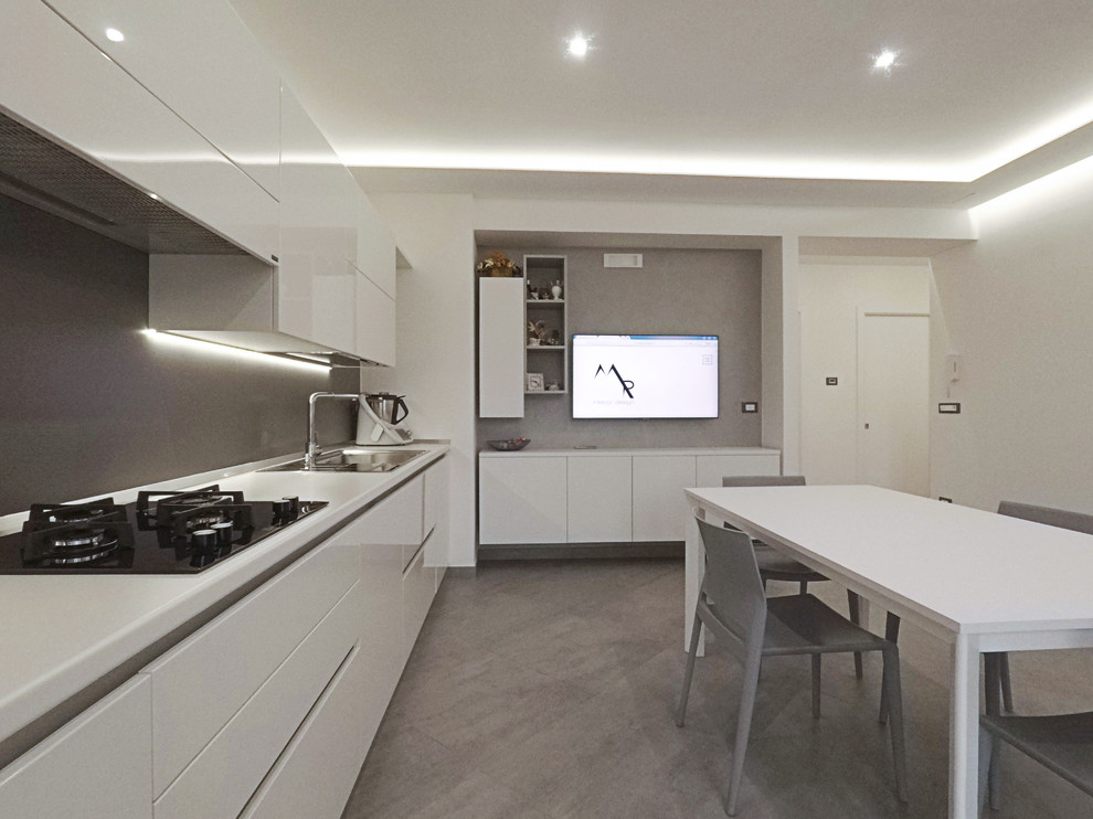 This is an example of a mid-sized contemporary single-wall open plan kitchen in Bari with a drop-in sink, flat-panel cabinets, white cabinets, solid surface benchtops, grey splashback, porcelain splashback, stainless steel appliances, porcelain floors, no island, grey floor and white benchtop.