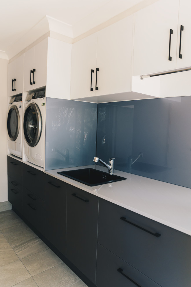 Photo of a large modern galley dedicated laundry room in Brisbane with a single-bowl sink, flat-panel cabinets, grey cabinets, quartz benchtops, grey splashback, glass sheet splashback, ceramic floors, a side-by-side washer and dryer, grey floor and grey benchtop.