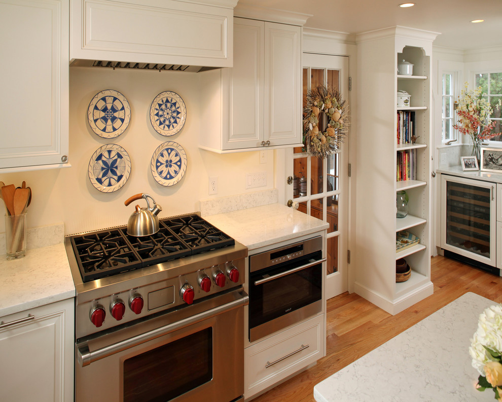Inspiration for a mid-sized transitional l-shaped medium tone wood floor eat-in kitchen remodel in Boston with white cabinets, an island, a farmhouse sink, raised-panel cabinets, marble countertops, white backsplash, glass sheet backsplash and stainless steel appliances