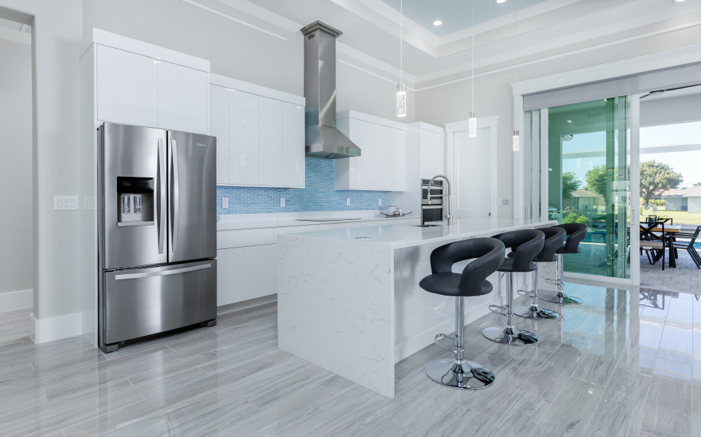 This is an example of a large mediterranean single-wall open plan kitchen in Miami with a farmhouse sink, flat-panel cabinets, white cabinets, quartz benchtops, blue splashback, ceramic splashback, stainless steel appliances, ceramic floors, with island, grey floor, white benchtop and recessed.