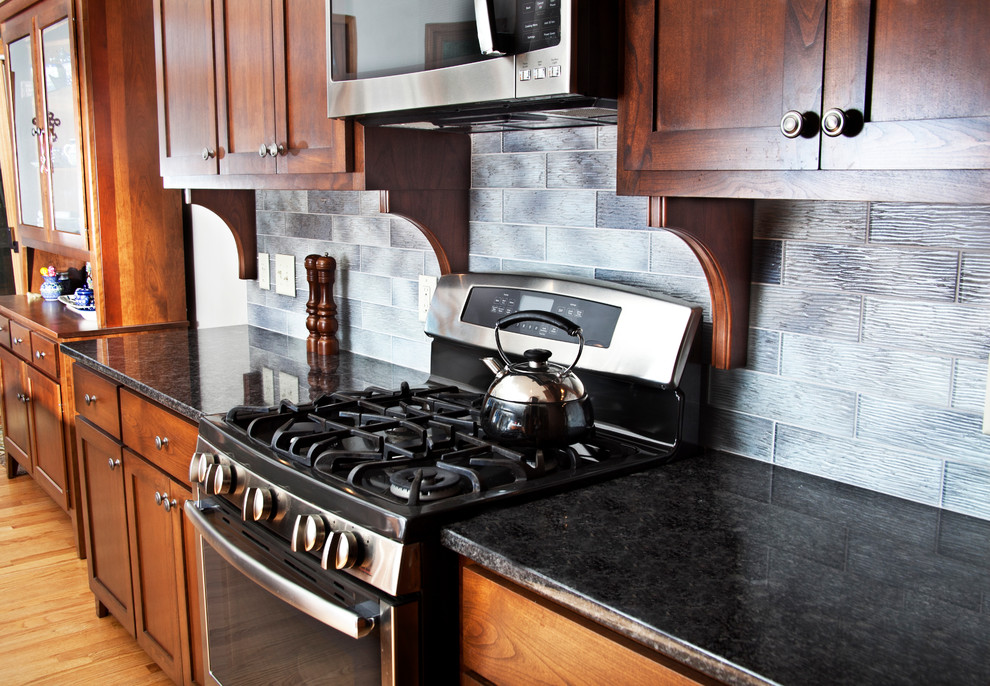 Transitional l-shaped light wood floor open concept kitchen photo in Milwaukee with a farmhouse sink, shaker cabinets, medium tone wood cabinets, granite countertops, gray backsplash, glass tile backsplash, stainless steel appliances and an island