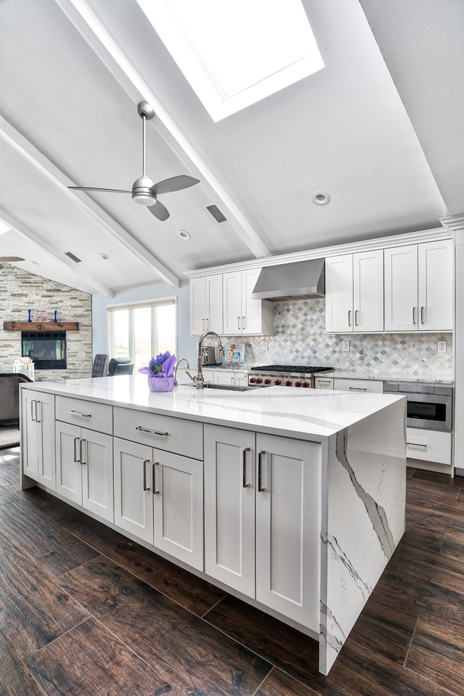 Photo of an expansive beach style galley eat-in kitchen in New York with a single-bowl sink, shaker cabinets, white cabinets, quartz benchtops, white splashback, marble splashback, stainless steel appliances, porcelain floors, multiple islands, brown floor and white benchtop.