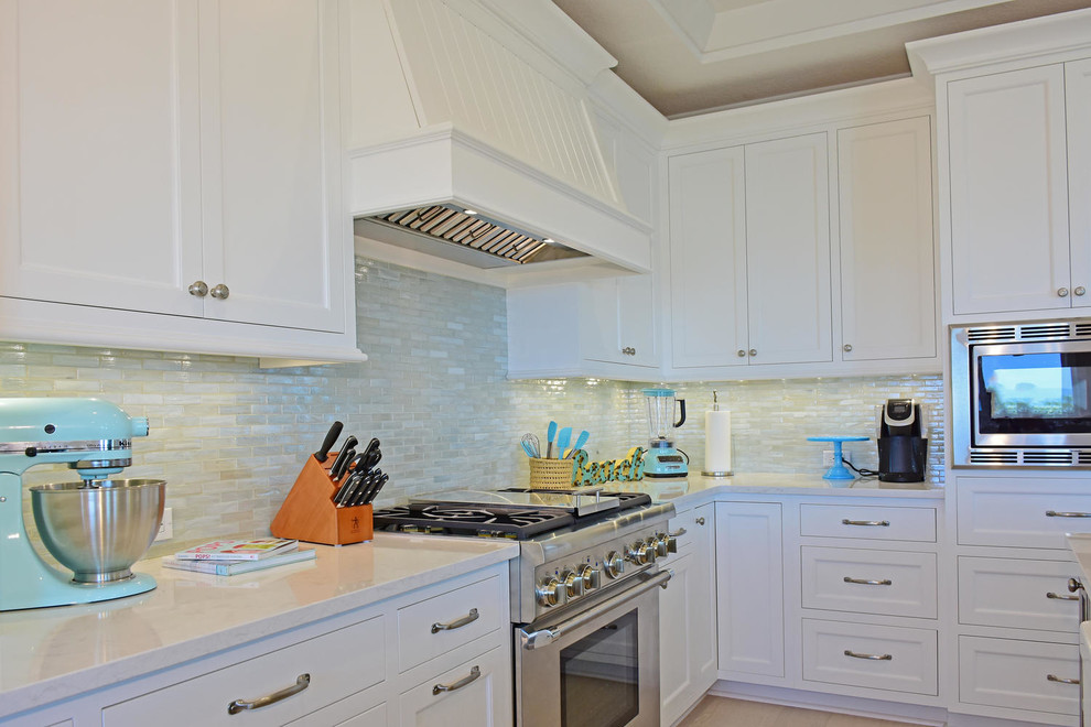 Inspiration for a mid-sized beach style single-wall eat-in kitchen in Tampa with a farmhouse sink, recessed-panel cabinets, white cabinets, beige splashback, glass tile splashback, stainless steel appliances, light hardwood floors and with island.