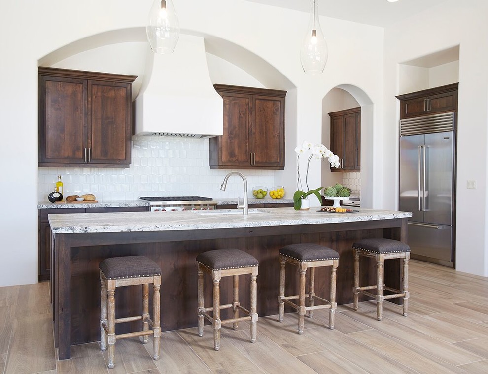 Example of a mid-sized transitional l-shaped medium tone wood floor and brown floor open concept kitchen design in Phoenix with an undermount sink, shaker cabinets, brown cabinets, marble countertops, white backsplash, porcelain backsplash, stainless steel appliances and an island