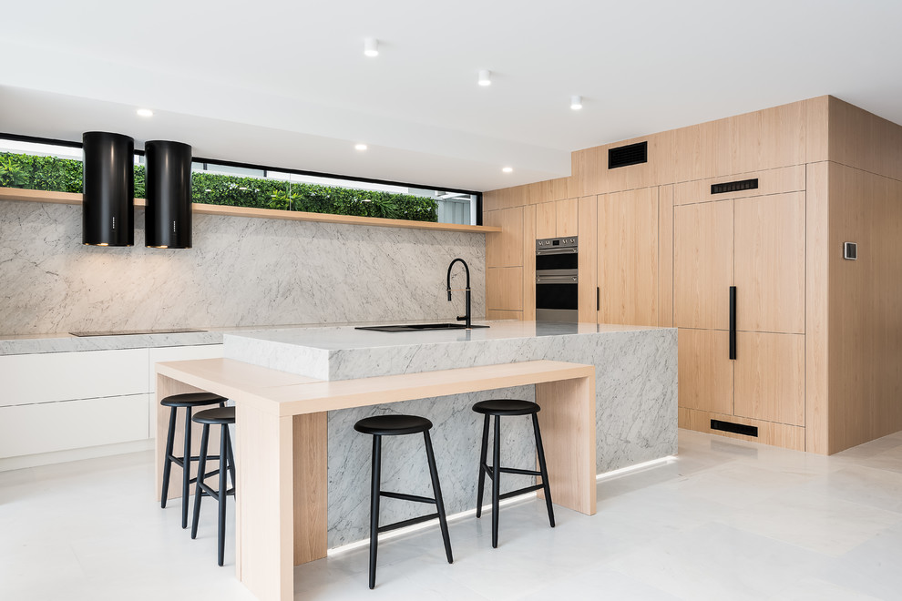 This is an example of a large modern l-shaped kitchen pantry in Brisbane with an undermount sink, beaded inset cabinets, light wood cabinets, marble benchtops, white splashback, marble splashback, black appliances, marble floors, with island, white floor and white benchtop.