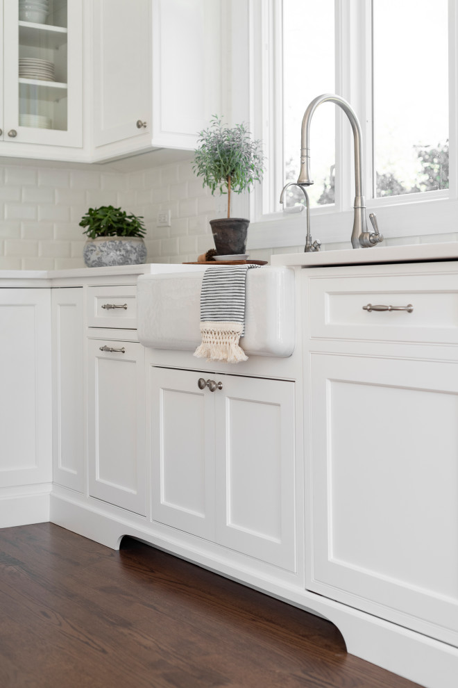 This is an example of a mid-sized transitional l-shaped eat-in kitchen in Chicago with a farmhouse sink, beaded inset cabinets, white cabinets, quartz benchtops, white splashback, ceramic splashback, panelled appliances, medium hardwood floors, with island, brown floor and white benchtop.