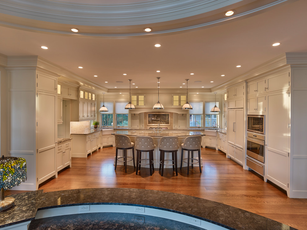Photo of an expansive traditional u-shaped eat-in kitchen in Other with a farmhouse sink, recessed-panel cabinets, white cabinets, limestone benchtops, beige splashback, travertine splashback, panelled appliances, dark hardwood floors, with island, brown floor and brown benchtop.