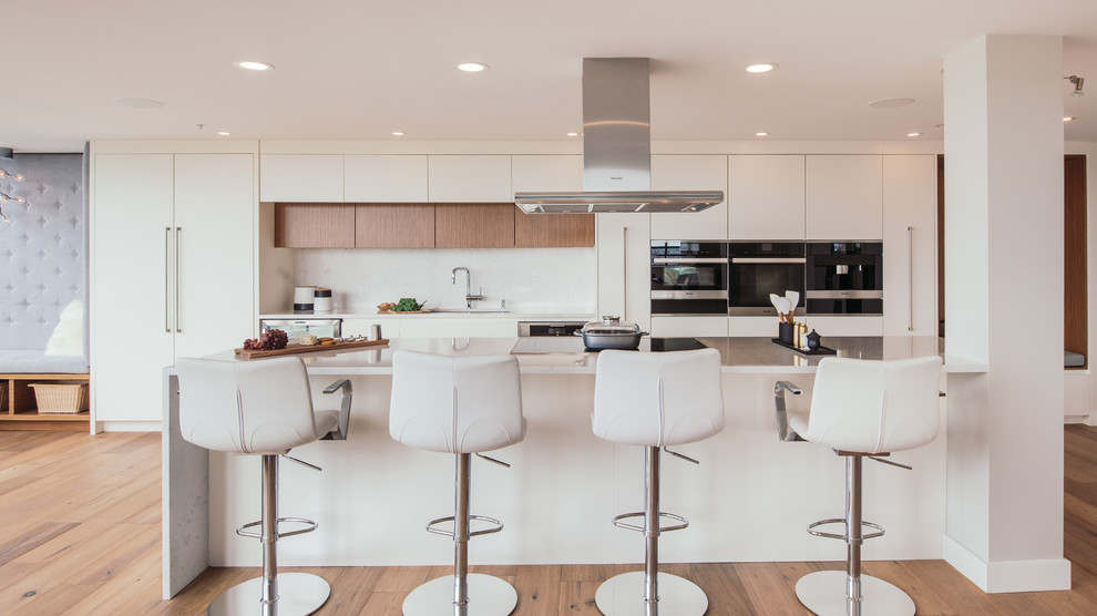 This is an example of a large contemporary galley open plan kitchen in Vancouver with a single-bowl sink, flat-panel cabinets, white cabinets, quartz benchtops, white splashback, stone slab splashback, panelled appliances, with island and medium hardwood floors.