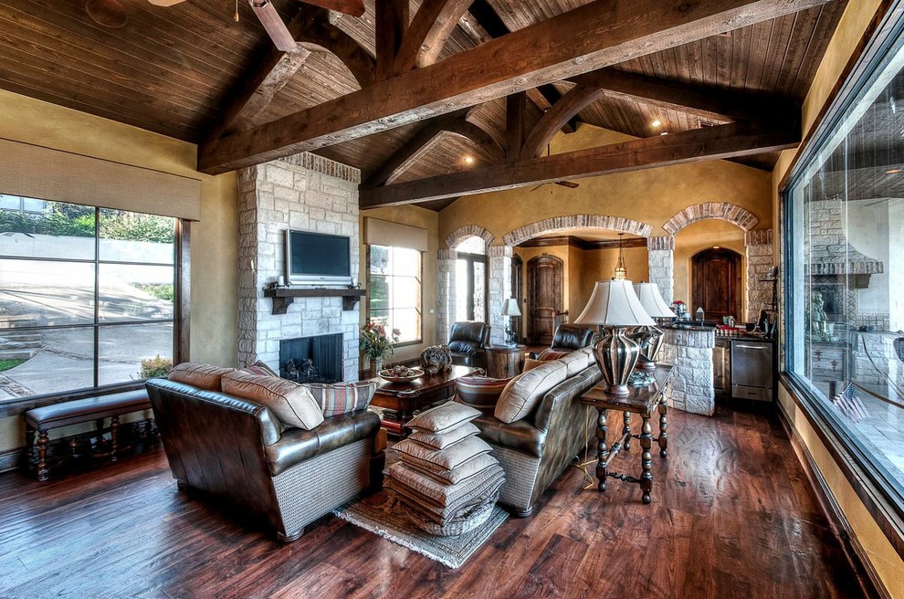 Large country open concept living room in Austin with beige walls, dark hardwood floors, a standard fireplace, a stone fireplace surround, a wall-mounted tv and brown floor.
