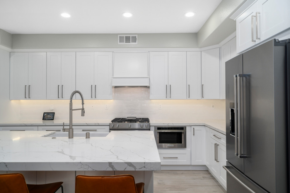 Kitchen - mid-sized transitional laminate floor and gray floor kitchen idea in Orange County with a farmhouse sink, shaker cabinets, white cabinets, white backsplash, subway tile backsplash, stainless steel appliances, an island and white countertops