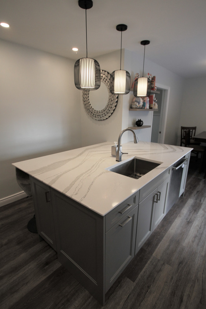 This is an example of a mid-sized contemporary l-shaped eat-in kitchen in Toronto with a single-bowl sink, recessed-panel cabinets, white cabinets, quartz benchtops, grey splashback, glass tile splashback, stainless steel appliances, laminate floors, with island, grey floor and white benchtop.