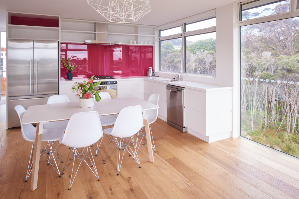 This is an example of a small contemporary single-wall eat-in kitchen in Auckland with a single-bowl sink, flat-panel cabinets, white cabinets, quartz benchtops, pink splashback, glass sheet splashback, stainless steel appliances, light hardwood floors and no island.