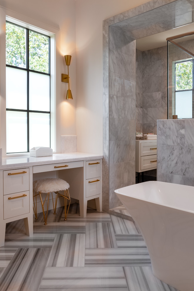 This is an example of a large transitional master bathroom in Houston with recessed-panel cabinets, white cabinets, a freestanding tub, a double shower, gray tile, white tile, limestone, white walls, limestone floors, an undermount sink, solid surface benchtops, white floor, a hinged shower door and white benchtops.