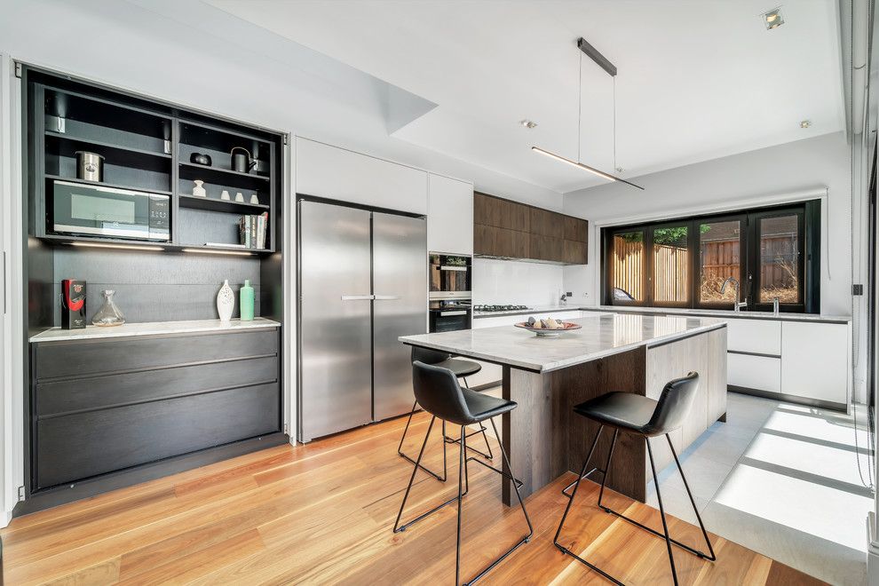 Large contemporary l-shaped eat-in kitchen in Sydney with a double-bowl sink, flat-panel cabinets, white cabinets, marble benchtops, white splashback, glass sheet splashback, stainless steel appliances, medium hardwood floors, with island, brown floor and grey benchtop.