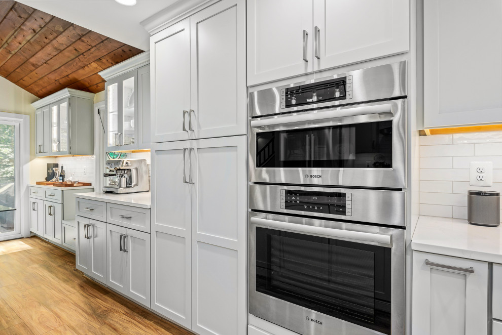 Large contemporary kitchen in DC Metro with a farmhouse sink, recessed-panel cabinets, white splashback, porcelain splashback, stainless steel appliances, medium hardwood floors, with island, brown floor and white benchtop.