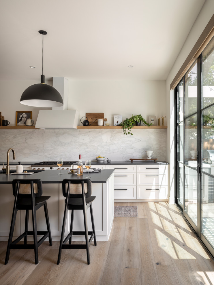 Large transitional l-shaped light wood floor open concept kitchen photo in Austin with an undermount sink, shaker cabinets, white cabinets, soapstone countertops, white backsplash, marble backsplash, white appliances, an island and black countertops