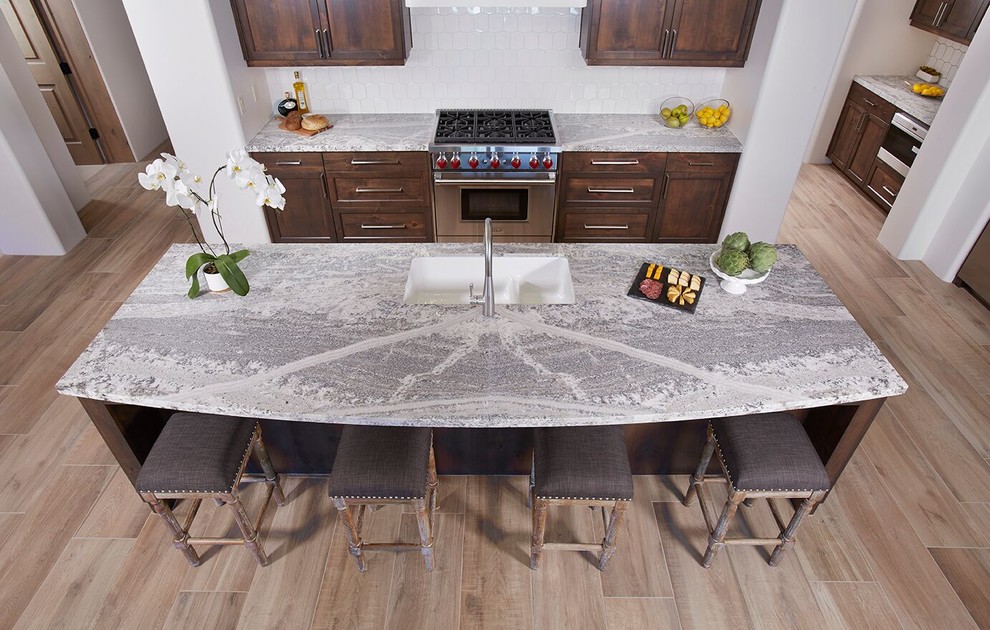 Open concept kitchen - mid-sized transitional l-shaped medium tone wood floor and brown floor open concept kitchen idea in Phoenix with an undermount sink, shaker cabinets, brown cabinets, marble countertops, white backsplash, porcelain backsplash, stainless steel appliances and an island
