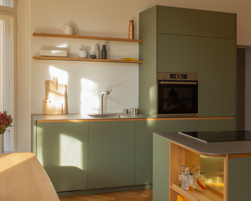 This is an example of a mid-sized contemporary galley open plan kitchen in Berlin with a single-bowl sink, flat-panel cabinets, green cabinets, stainless steel benchtops, white splashback, stainless steel appliances, medium hardwood floors, with island and grey benchtop.