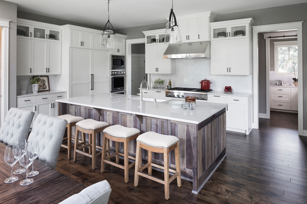 Photo of a large traditional l-shaped eat-in kitchen in Minneapolis with a farmhouse sink, recessed-panel cabinets, white cabinets, quartz benchtops, white splashback, ceramic splashback, stainless steel appliances, dark hardwood floors, with island and brown floor.