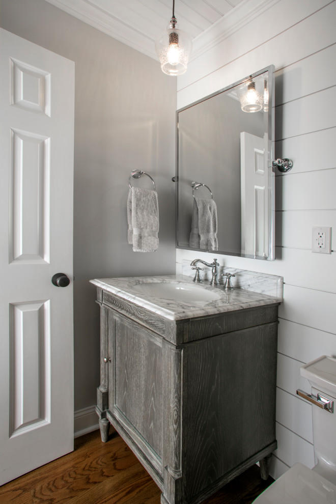 Photo of a mid-sized eclectic powder room in Philadelphia with furniture-like cabinets, grey cabinets, a one-piece toilet, grey walls, medium hardwood floors, an undermount sink, marble benchtops, brown floor, white benchtops, a freestanding vanity, wood and planked wall panelling.