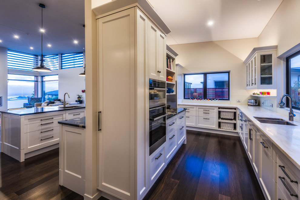 Large traditional u-shaped kitchen pantry in Christchurch with an undermount sink, shaker cabinets, white cabinets, granite benchtops, white splashback, glass sheet splashback, stainless steel appliances, dark hardwood floors, with island, brown floor and black benchtop.