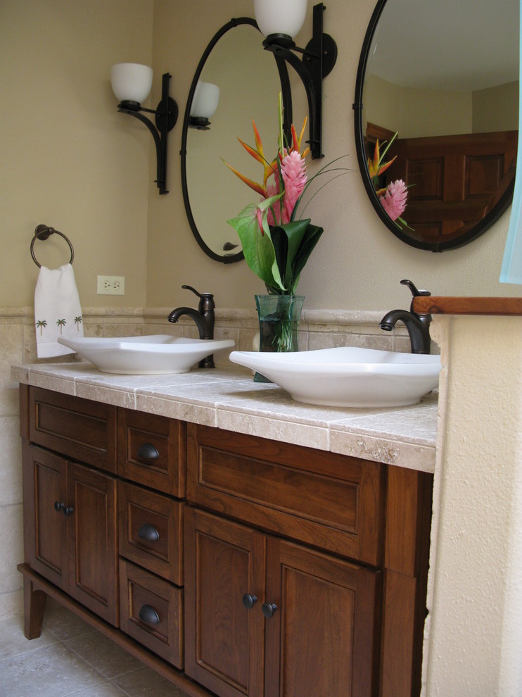 Photo of a mid-sized tropical bathroom in Seattle with a vessel sink, dark wood cabinets, tile benchtops, an alcove tub, a shower/bathtub combo, a two-piece toilet, beige tile, stone tile, beige walls, travertine floors and recessed-panel cabinets.