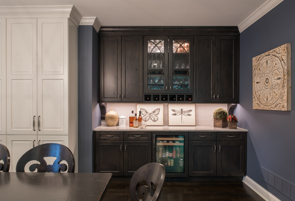This is an example of a mid-sized transitional single-wall wet bar in Minneapolis with no sink, recessed-panel cabinets, dark wood cabinets, granite benchtops, white splashback, subway tile splashback, dark hardwood floors and brown floor.