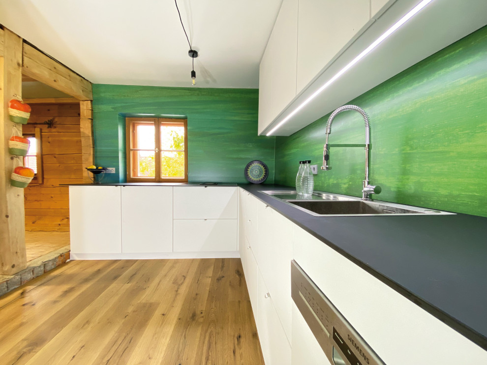 Küche ohne Insel in L-Form mit flächenbündigen Schrankfronten, weißen Schränken, Küchenrückwand in Grün, hellem Holzboden, schwarzer Arbeitsplatte, Doppelwaschbecken, Mineralwerkstoff-Arbeitsplatte und Elektrogeräten mit Frontblende in Munich