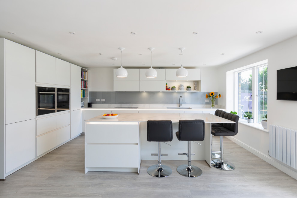 Photo of a medium sized contemporary l-shaped kitchen/diner in Gloucestershire with flat-panel cabinets, white cabinets, engineered stone countertops, grey splashback, glass sheet splashback, black appliances, an island and white worktops.