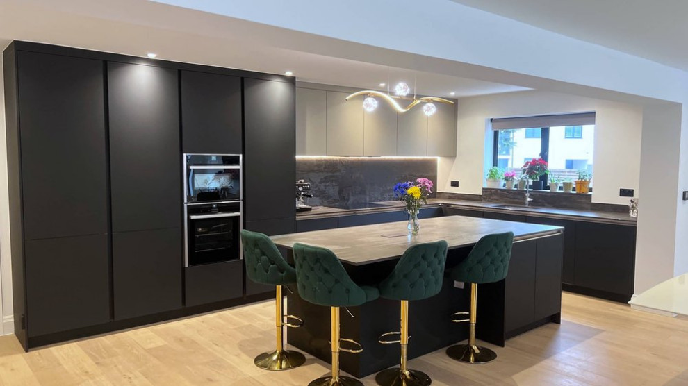 Photo of a medium sized contemporary l-shaped open plan kitchen in London with a single-bowl sink, flat-panel cabinets, black cabinets, composite countertops, black splashback, black appliances, light hardwood flooring, an island, brown floors and multicoloured worktops.