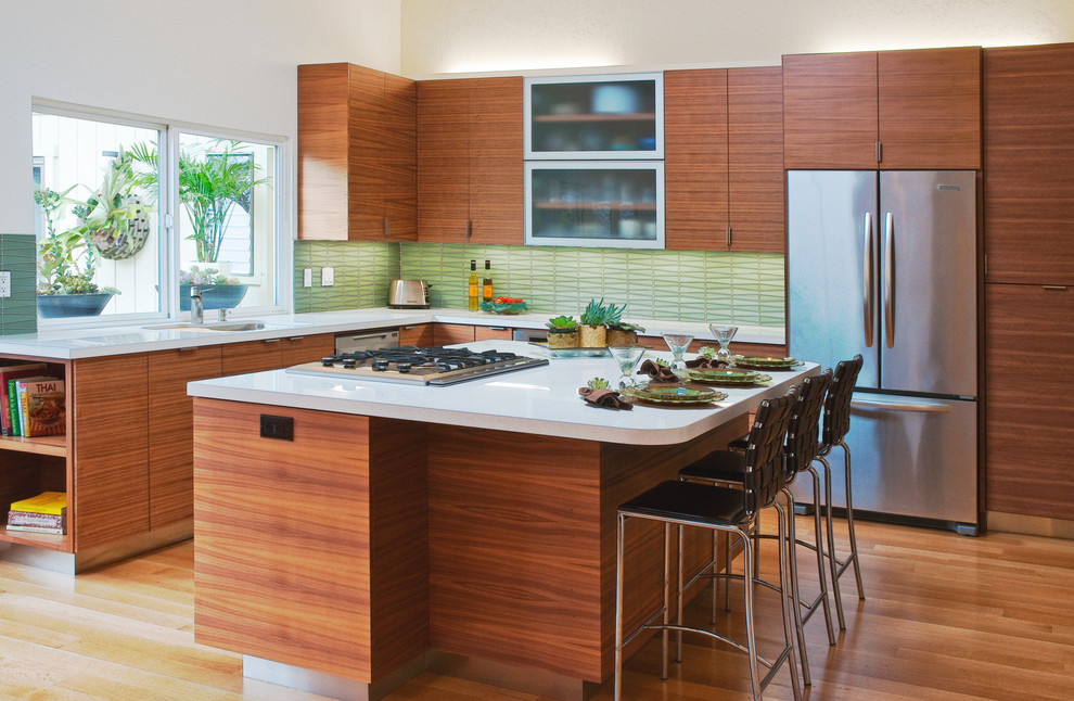 This is an example of a midcentury l-shaped eat-in kitchen in San Francisco with stainless steel appliances, flat-panel cabinets, medium wood cabinets, green splashback, an undermount sink, solid surface benchtops, light hardwood floors, with island and glass tile splashback.