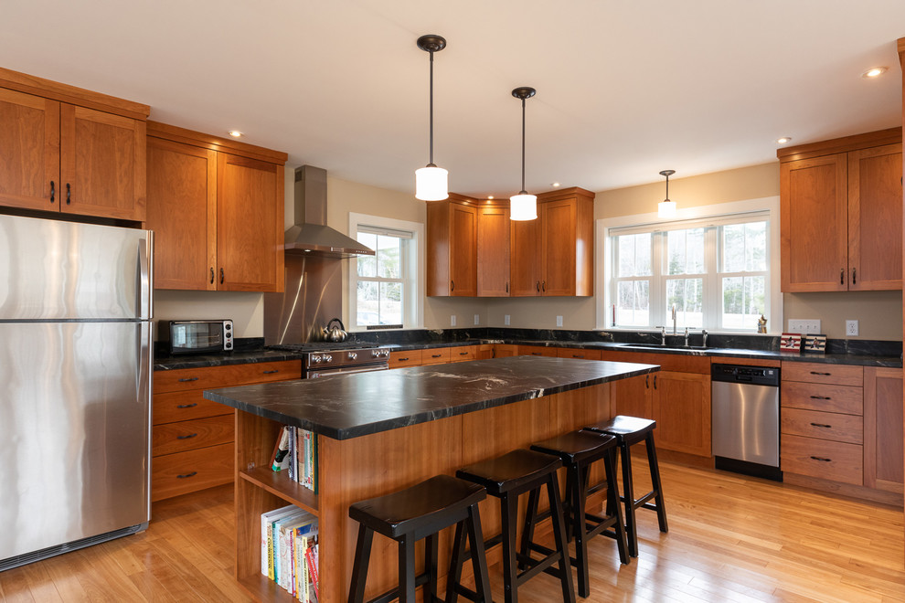 Photo of a large country l-shaped eat-in kitchen in Portland Maine with an undermount sink, shaker cabinets, medium wood cabinets, granite benchtops, stainless steel appliances, light hardwood floors, with island, brown floor and black benchtop.