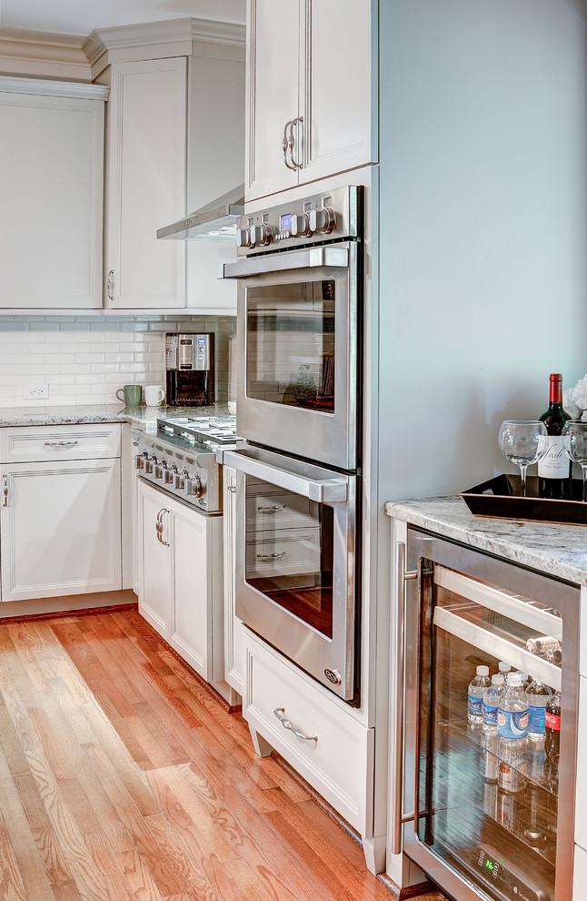 Example of a mid-sized classic u-shaped medium tone wood floor kitchen design with a farmhouse sink, beaded inset cabinets, gray cabinets, granite countertops, white backsplash, porcelain backsplash, stainless steel appliances and an island
